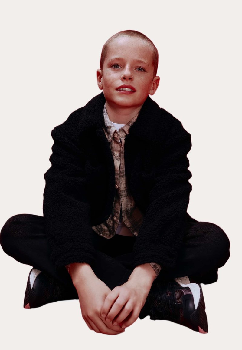 Sort, fleece-fórret jakke over en ternet skjorte, sorte bukser og sorte sneakers. Siddeposition med hænderne foldet på knæene. Neutral baggrund.