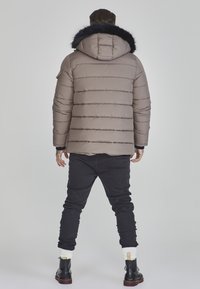 Beige pufferjakke med aftagelig sort faux fur-hætte, quiltet design og ribbede manchetter, parret med sorte jeans og sorte støvler.