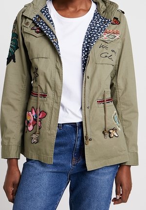 Personne portant une veste vert olive avec des broderies florales et d’insectes, une chemise blanche et un jean bleu, les mains reposant le long du corps.