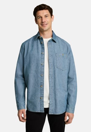 Mann trägt ein offenes hellblaues Hemd über einem weißen T-Shirt, steht vor einem einfarbigen Hintergrund und lächelt leicht.