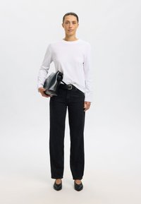 Schwarze Jeans mit geradem Bein, kombiniert mit einem langärmligen weißen Hemd, hält eine schwarze Ledertasche. Einfaches Design, keine sichtbaren Muster oder Akzente.