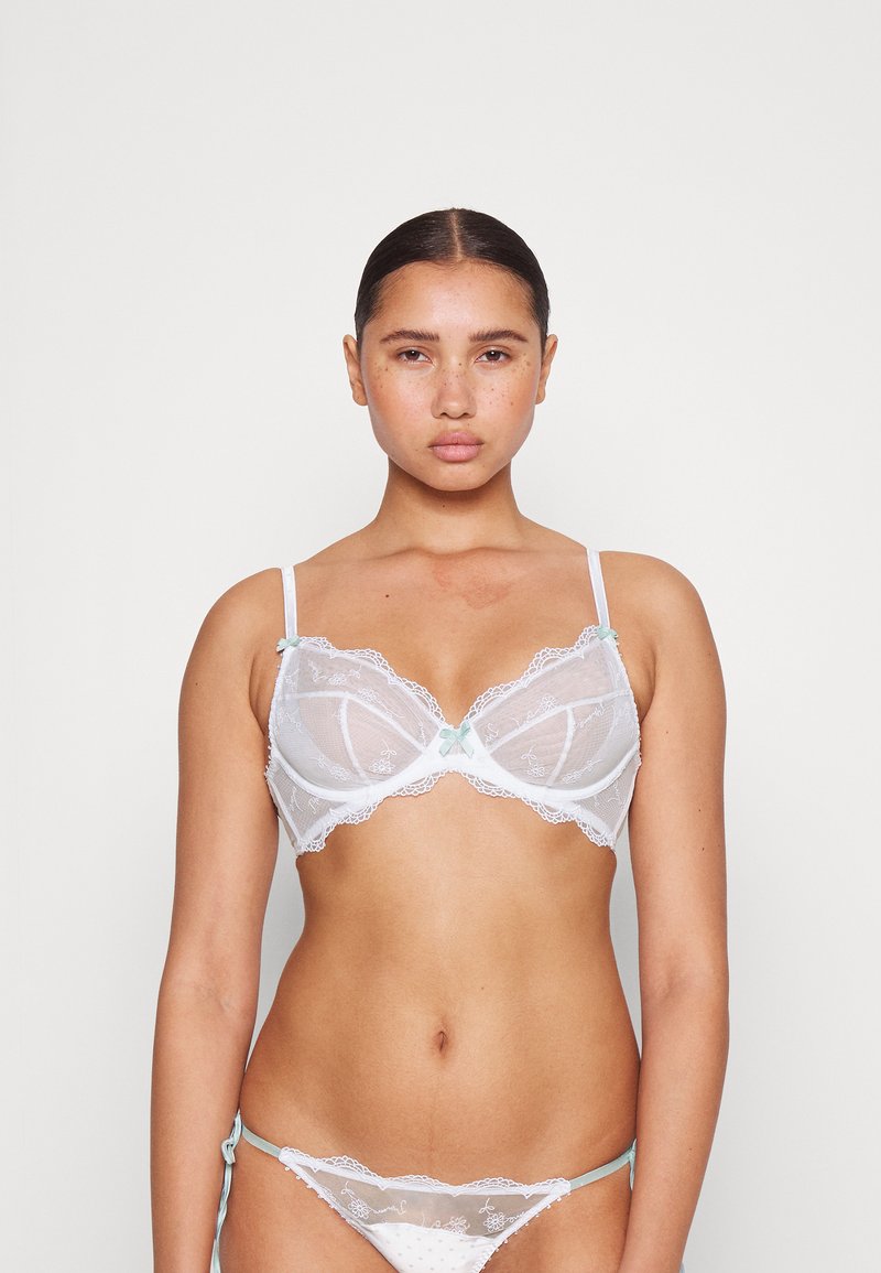Sutiã de renda branca com padrões florais bordados, copas suaves, alças ajustáveis e detalhe de laço azul claro ao centro. Usado com parte de baixo combinando.