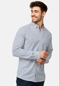 Weißes Hemd mit blauen geometrischen Mustern, langen Ärmeln und klassischem Kragen. Hat einen abgerundeten Saum und geknöpfte Manschetten.