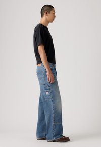 Camiseta negra de manga corta combinada con jeans de mezclilla azul claro de pierna ancha que cuentan con múltiples bolsillos y una etiqueta de parche en la parte trasera. Zapatos marrones.