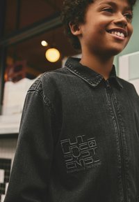 Svart denimjacka med dragkedja framtill, krage och texturerad broderi med "FUTURE SYSTEMS" på bröstet.
