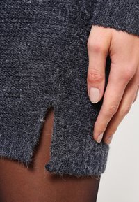 Gray textured sweater with a visible slit; hand resting gently on the fabric, revealing an intricate knit pattern and soft finish.