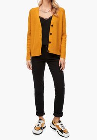 Senapsgul ribbad cardigan med svarta knappar, lager på lager över en svart topp med spetskant, kombinerad med svarta byxor och flersfärgade sneakers.