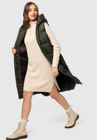Langer, dunkelgrüner Steppweste mit Kapuze, offen über einem cremefarbenen Strickkleid mit seitlichen Schlitzen getragen, kombiniert mit beigen Schnürstiefeln.