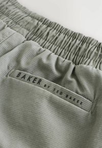 Elastische tailleband in zacht olijfgroen stof met fijne ribbels; voorzien van een zwart label met "BAKER by TED BAKER" erop gestikt.