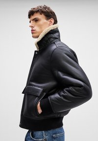 Black leather bomber jacket with a plush beige collar, quilted texture at the neck, and a front patch pocket. Ribbed cuffs and hem.