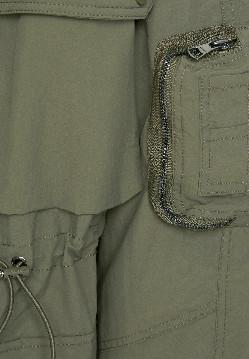 Veste olive en tissu texturé, dotée d'une poche zippée, d'une taille à cordon de serrage et de détails de couture visibles. Design utilitaire moderne.