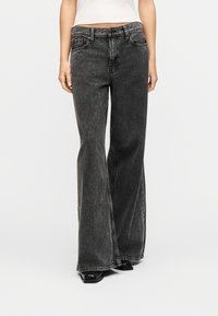 Γυναίκα που φοράει ψηλόμεσα, φαρδιά ξεθωριασμένα μαύρα τζιν, συνδυασμένα με λευκό κοντό τοπ και μαύρα παπούτσια, στέκεται μπροστά σε λευκό φόντο.