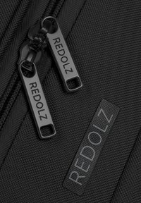 Gros plan d’un sac en tissu noir avec deux curseurs de fermeture éclair noirs et une étiquette, tous marqués du nom "REDOLZ" en lettres majuscules.