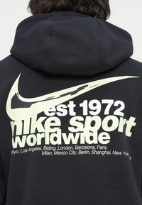 Persona che indossa un hoodie nero con un grande logo Nike di un pallido giallo e la scritta "est 1972 Nike Sport Worldwide" con i nomi delle città stampati sul retro.