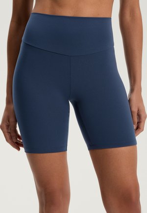 Vrouw draagt donkerblauwe, hooggetailleerde, nauwsluitende fietsbroek, getoond van midden torso tot midden dij, staand met armen ontspannen langs het lichaam.