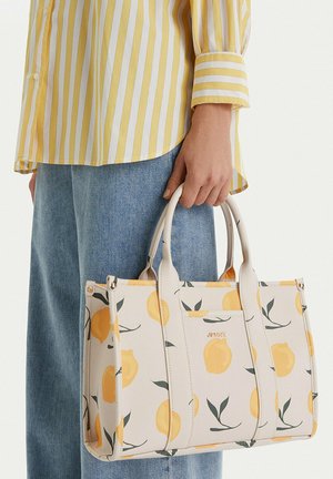 Persona che tiene una borsa a tracolla beige con stampa di limoni gialli, indossa una maglietta a righe gialle e bianche e jeans blu, su sfondo semplice.