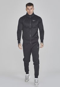 Chándal deportivo negro confeccionado en tejido suave, que incluye una chaqueta con cremallera y cuello alto, así como pantalones ajustados. Con acento de un logo.
