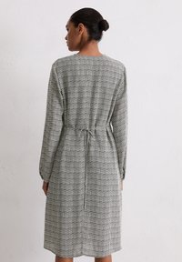 Frau mit einem Dutt, die ein knielanges, langärmliges Kleid mit einem schwarz-weißen geometrischen Muster trägt, mit dem Rücken zu einer weißen Wand stehend.