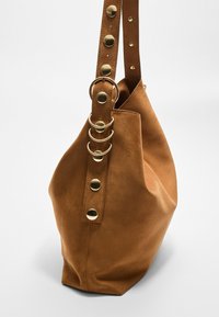 Borsa a spalla in suede marrone con una forma morbida, dotata di una tracolla larga adornata da borchie dorate e anelli circolari per i dettagli in metallo.