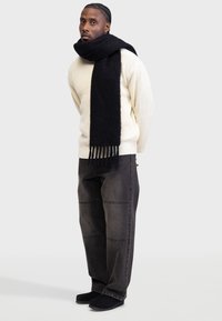 Bufanda negra de textura de punto, llevada con un suéter crema de punto trenzado. Jeans anchos gris oscuro con detalles de costura. Zapatos negros tipo slip-on.