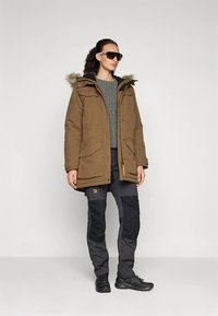 Barna parka szőrme béléssel ellátott kapucnival, szürke pulóver, fekete nadrág térdpárnákkal és fekete lábbeli; a modell napszemüveget visel.