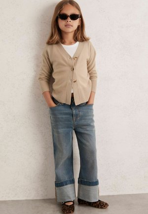 Beige strikket cardigan med tre knapper over en hvid top, parret med bredbenede denimjeans og leopardmønstrede flade sko. Solbriller fuldender looket.