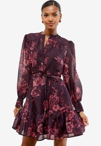 Donna che indossa un vestito a maniche lunghe floreale di un viola scuro con orlo arricciato e cintura in vita, in piedi contro uno sfondo bianco semplice.