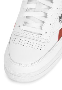 Adidași albi cu partea superioară din piele netedă, accente roșii, cutie de deget perforată, șireturi albe plate și talpă din cauciuc texturată cu puncte ridicate.