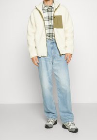 Man draagt een crèmekleurig fleecejack met olijfgroene zak, geruit overhemd, lichtblauwe spijkerbroek en wit-grijze wandelschoenen, staand op een grijze vloer.