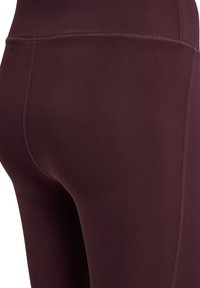 Burgundowe legginsy o gładkiej fakturze, wysokim pasie i szwach stworzonych płaską nitką, ukazujące dopasowany kształt i brak widocznych wzorów czy metalowych elementów.