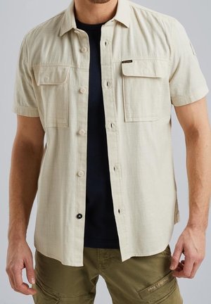 Homme portant une chemise beige à manches courtes boutonnée par-dessus un t-shirt noir et un pantalon cargo vert olive avec un patch de marque sur la poche cuisse.