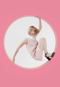 Model in een roze satijnen slipdress met een witte kraag, gekleed in beige panty’s en zwarte patenten schoenen, ingekaderd in een ronde witte cirkel tegen een roze achtergrond.