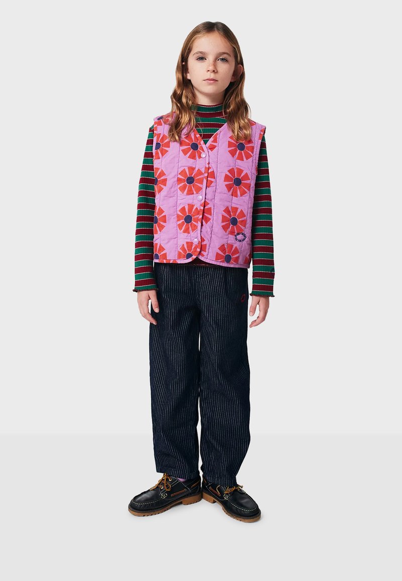 Gepolsterte pinke Weste mit rotem Blumenmuster über einem gestreiften grünen und schwarzen Langarmhemd, kombiniert mit dunkel gestreiften Hosen und schwarzen Schuhen.