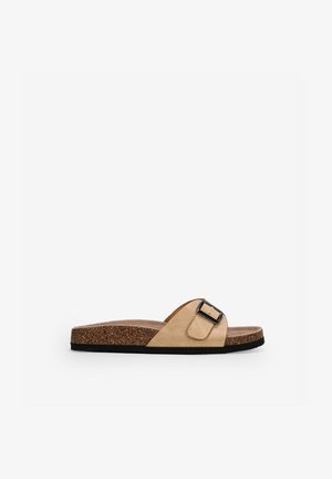Sandalia tipo chancleta color beige con una correa ancha que tiene una hebilla negra, plantilla de corcho y suela de goma negra sobre un fondo blanco.