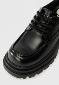 Melna ādas zamšota kurpe ar masīvu zoli, kurai ir teksturēts virspusē, noapaļots purngals un izteikta šuvju detaļa.