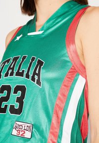 Zelený basketbalový dres s černým textem "ITALIA 23", červenými bočními akcenty a bílými pruhy. Vyrobeno z lesklé látky se záplatou s nápisem "JADED LDN 92."
