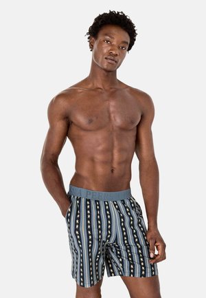 Homme torse nu debout, une main dans la poche, portant un boxer rayé avec des motifs d'étoiles et une ceinture de marque.