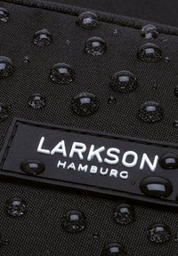 Närbild av svart texturerad tyg med vattendroppar och en rektangulär etikett med texten "LARKSON HAMBURG" i vita bokstäver.