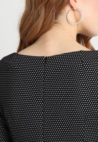 Tissu noir à motifs avec de petits carrés blancs, présentant un col rond et un détail de fermeture éclair au dos. Une grande boucle d'oreille en argent est visible.