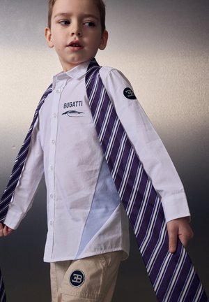 Junger Junge trägt ein weißes Bugatti-Shirt und beige Hose, hält einen langen lila-weiß gestreiften Schal über den Schultern.