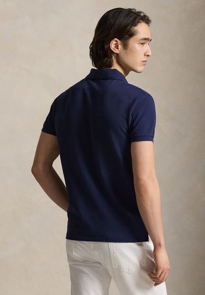 Μπλε ναυτικό πόλο πουκάμισο από υφασμάτινη υφή, με κοντά μανίκια και κλασικό γιακά. Φορεμένο με λευκό παντελόνι, εμφανίζεται από πίσω.