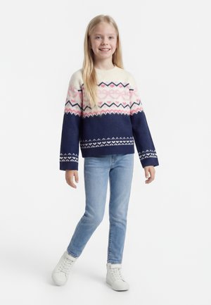 Ung jente står, iført en mønstret genser i marineblått, rosa og hvitt, lyseblå jeans og hvite høye joggesko, smilende.