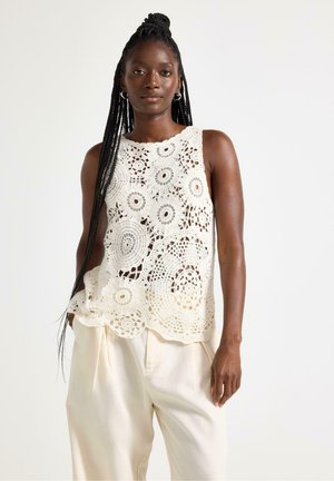 Kvinde med langt flettet hår iført en hvid ærmeløs hækletrøje og løstsiddende cremefarvede bukser mod en ensfarvet hvid baggrund.