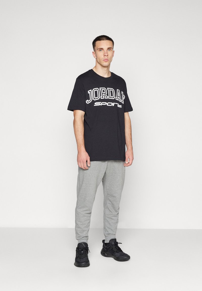 Camiseta de algodón negra con el texto blanco "JORDAN" y "sport", combinada con pantalones de chándal gris claro. El modelo lleva zapatillas deportivas negras.