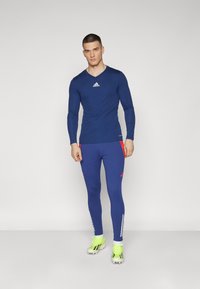 Blå långärmad träningsskjorta med V-ringning, Adidas-logotyp på bröstet; i kombination med blå leggings med röda accenter och vita ränder.
