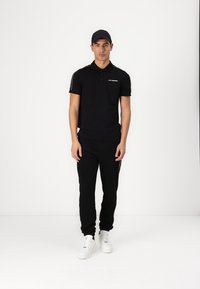 Svart polo med korta ärmar, som har en logotyp och guld detaljer, kombinerad med svarta träningbyxor och vita sneakers. Enkel, atletisk design.