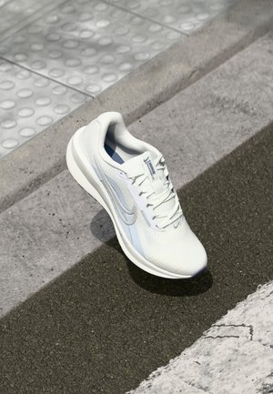 Chaussure de course Nike blanche avec des accents gris clair placée au bord d'un trottoir texturé et d'une route asphaltée.