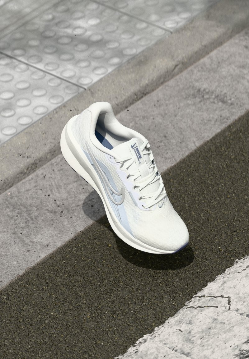 Chaussure de course Nike blanche avec des accents gris clair placée au bord d'un trottoir texturé et d'une route asphaltée.