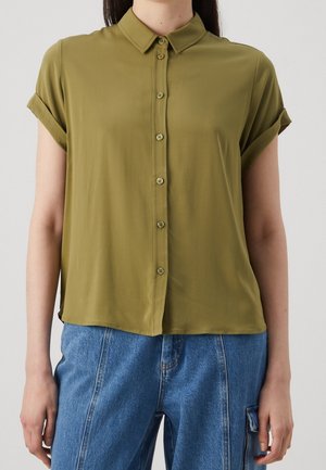 Femme portant une chemise olive à manches courtes avec boutons et poignets retroussés, ainsi qu'un pantalon en denim bleu avec des détails de poches latérales.