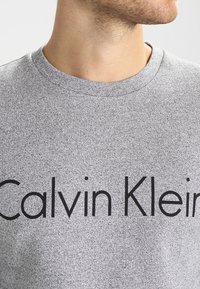 Sudadera gris con un acabado texturizado, que presenta un destacado logo negro "Calvin Klein" en la parte frontal en una fuente en negrita.